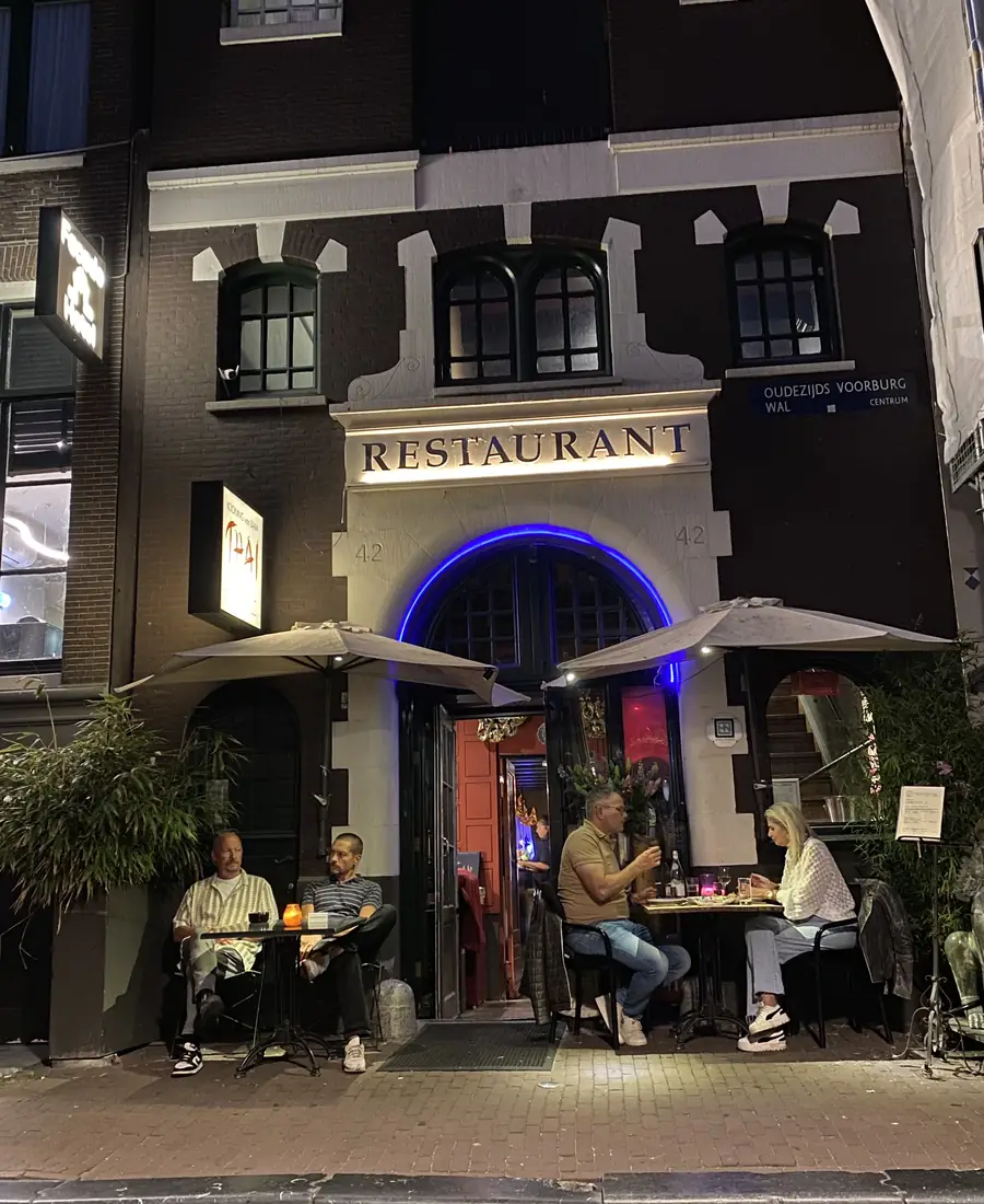 De gevel van het restaurant aan de Oudezijds Voorburgwal in de avond, met zitplekken buiten
